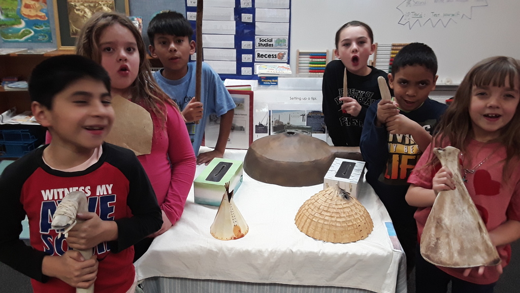 Students holding Native American tools, standing around a display of Indian Homes in Kansas models.